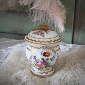 Könnte beinhalten: Eine weiße Porzellandose mit Deckel, verziert mit handgemalten Blumenmustern in Rosa, Rot, Lila und Blau. Die Dose und der Deckel sind mit goldenen Akzenten versehen. Der Deckel ist mit einem goldfarbenen, blütenförmigen Knauf versehen.