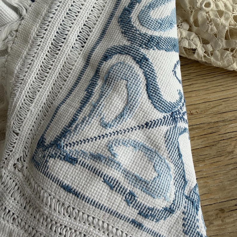 Puede incluir: Textil blanco y azul estampado. La tela presenta una textura de tejido de gofre con dise&ntilde;os azules bordados. El borde tiene un detalle de flecos. El textil es probablemente una manta, un coj&iacute;n o un pa&ntilde;o decorativo.