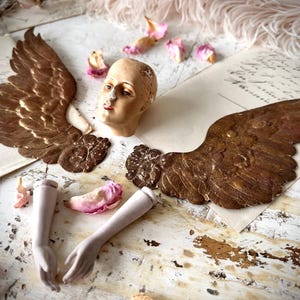May include: A vintage angel figurine in pieces, featuring bronze wings, a pale head with red lips, and detached arms. The components are arranged on aged paper, with scattered pink flower petals.