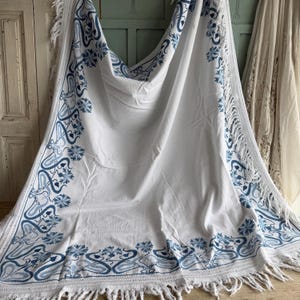 Puede incluir: Textil blanco con bordado floral y de volutas azules. Los bordes est&aacute;n acabados con flecos. El dise&ntilde;o presenta un patr&oacute;n repetitivo de flores estilizadas y l&iacute;neas fluidas, creando un borde decorativo.