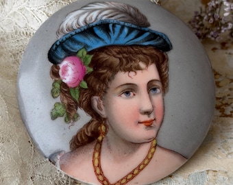 Puede incluir: Una placa de cerámica redonda con un retrato pintado a mano de una persona con cabello castaño rizado, que lleva un sombrero azul con una pluma blanca y una rosa rosa. La figura lleva un collar de cadena de tono dorado sobre un fondo gris claro.