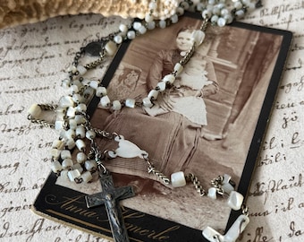 Puede incluir: Un rosario vintage con cuentas blancas y cruces plateadas descansa sobre una fotografía en tonos sepia. La fotografía presenta a una mujer sosteniendo a un niño. El rosario está drapeado sobre la foto, creando un efecto en capas.