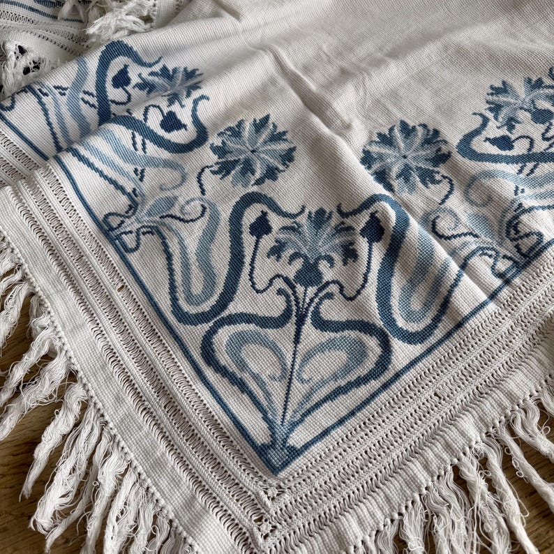 Puede incluir: Textil color crema con un estampado floral azul y flecos. El dise&ntilde;o presenta flores estilizadas y l&iacute;neas fluidas en tonos azules. Los bordes est&aacute;n acabados con un borde de ganchillo y flecos largos.