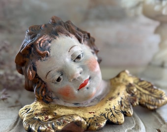 Puede incluir: Una placa de pared de cerámica con forma de querubín, con rostro pintado a mano, mejillas sonrosadas, cabello castaño y labios rojos brillantes. La figura presenta un cuello dorado texturizado que recuerda a hojas estilizadas.