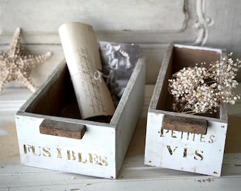 Puede incluir: Dos cajas de almacenamiento de madera blanca con asas marrones y texto con plantilla. Una caja contiene un documento enrollado y una fotografía, la otra flores secas. Una estrella de mar está en el fondo.