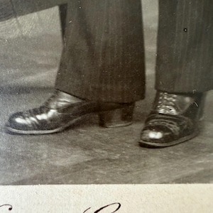 Può includere: Fotografia in bianco e nero di due paia di scarpe da uomo e della parte inferiore di due pantaloni gessati. Un paio di scarpe è a slip-on, l'altro è allacciato. La fotografia è firmata in basso.