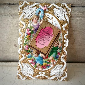 Pode incluir: Um cartão de felicitações vintage com uma moldura de filigrana dourada e uma borda de renda branca. O cartão apresenta figuras de querubins, flores e um livro com texto em alemão. A paleta de cores inclui dourado, branco, rosa, azul e verde.