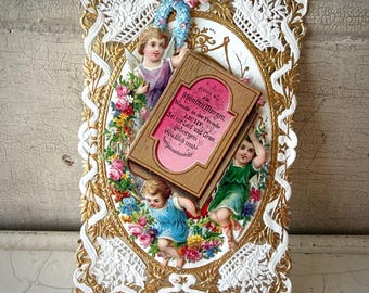 Puede incluir: Una tarjeta de felicitación vintage con un marco de filigrana dorado y un borde de encaje blanco. La tarjeta presenta figuras de querubines, flores y un libro con texto en alemán. La paleta de colores incluye dorado, blanco, rosa, azul y verde.