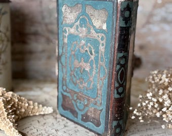Puede incluir: Un libro antiguo con una cubierta turquesa y detalles dorados ornamentados. La cubierta presenta un diseño central dentro de un marco decorativo. El lomo es marrón oscuro con letras doradas. El libro está colocado sobre un fondo de papel vintage.