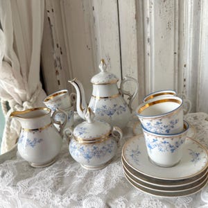 Könnte beinhalten: Vintage Porzellan-Teeservice mit Teekanne, Sahnekännchen, Zuckerdose, Tassen und Untertassen. Das Set hat eine weiße Basis mit blauen Blumenmustern und goldenen Verzierungen. Das Teeservice ist auf einer weißen Spitzendecke arrangiert.