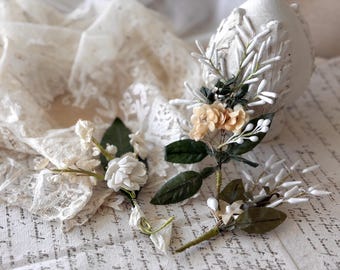 Puede incluir: Primer plano de boutonnieres florales con flores color crema, hojas verdes y detalles blancos. Los boutonnieres están dispuestos sobre una tela de encaje vintage y un fondo de escritura manuscrita.