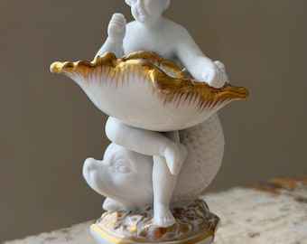 Puede incluir: Una figura de porcelana blanca que muestra a un querubín sentado sobre un delfín, sosteniendo un cuenco grande con forma de concha. La pieza presenta detalles en oro en los bordes de la concha, la base y los relieves en la parte inferior.