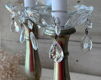 Puede incluir: Dos candelabros antiguos con adornos de cristal transparente. Los candelabros tienen una base de metal plateado y una parte superior en forma de cuenco de cristal transparente. Los cristales son en forma de lágrima y facetados.