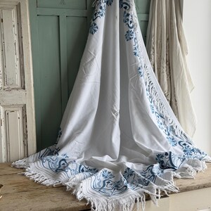 Puede incluir: Textil blanco con dise&ntilde;o floral y de volutas azules. La tela tiene un borde con flecos y est&aacute; drapeada sobre una superficie de madera. El patr&oacute;n azul se repite a lo largo de los bordes y en el centro del textil.