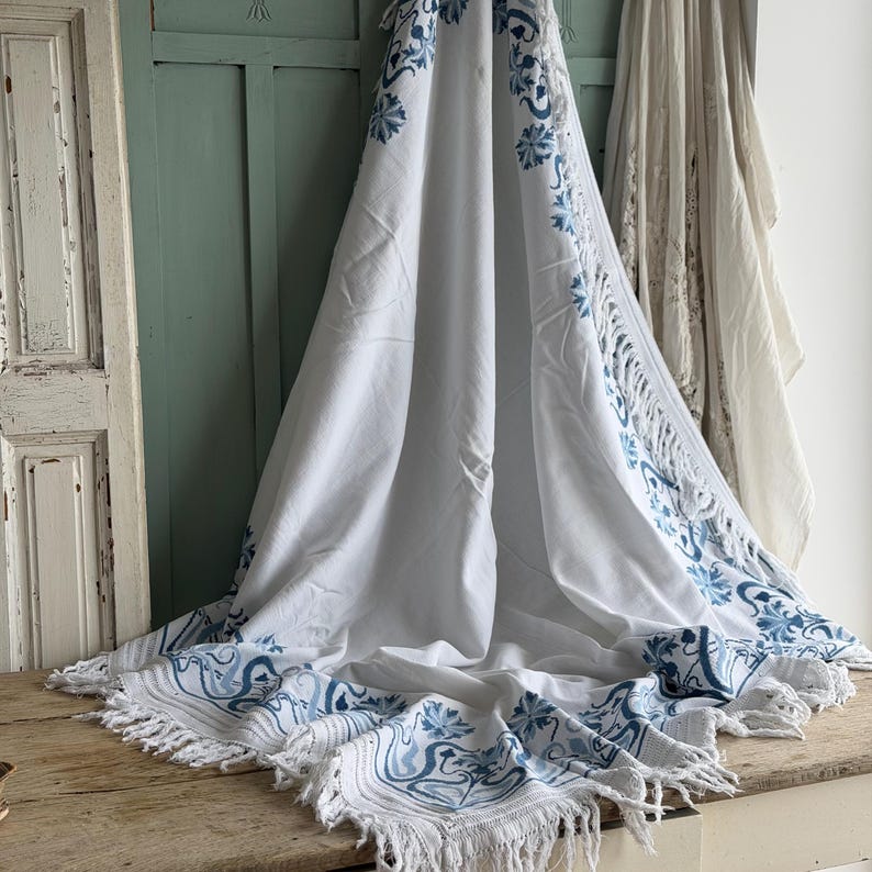 Puede incluir: Textil blanco con dise&ntilde;o floral y de volutas azules. La tela tiene un borde con flecos y est&aacute; drapeada sobre una superficie de madera. El patr&oacute;n azul se repite a lo largo de los bordes y en el centro del textil.