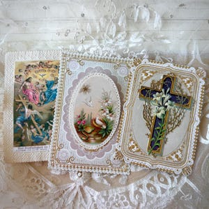 Pode incluir: Três cartões religiosos vintage com bordas ornamentadas. Um cartão retrata uma cruz com lírios, outro apresenta uma imagem oval de pombas e flores, e o terceiro mostra uma cena religiosa com figuras. Os cartões estão em um fundo de renda.