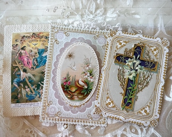 Puede incluir: Tres tarjetas religiosas vintage con bordes ornamentados. Una tarjeta representa una cruz con lirios, otra presenta una imagen ovalada de palomas y flores, y la tercera muestra una escena religiosa con figuras. Las tarjetas están sobre un fondo de encaje.