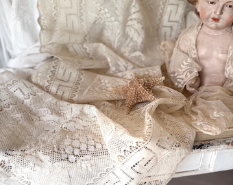 Puede incluir: Tela de encaje antiguo color crema con intrincados patrones florales y geométricos. Una muñeca vintage y una delicada estrella de mar acentúan el encaje drapeado. El encaje se exhibe sobre una superficie blanca.