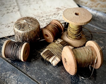 Puede incluir: Varias bobinas de hilo vintage en tonos dorados, marrones y bronce. Las bobinas de madera están dispuestas sobre una superficie oscura y texturizada, evocando la artesanía y el pasado.