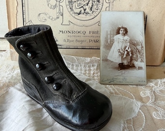 Puede incluir: Bota de bebé negra antigua con cierres de botón. La bota tiene una puntera redondeada y un tacón pequeño. Una fotografía vintage de un niño está en el fondo, junto con un libro vintage decorativo.
