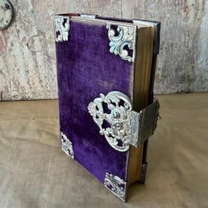 Può includere: Un libro vintage con una copertina in velluto viola scuro e protezioni angolari e chiusura in metallo argentato ornate. I bordi del libro sono dorati e la chiusura è finemente decorata con dettagli floreali e a volute. Il libro è chiuso.