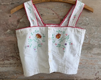 Puede incluir: Un top corto blanco sin mangas con cierre de botones frontal y patrones florales bordados en naranja, rosa y verde. La prenda está acentuada con un borde festoneado rojo a lo largo del escote y los tirantes, colgada en una percha de madera.