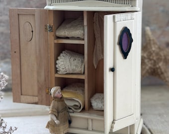 Puede incluir: Un armario de madera en miniatura pintado de color blanco roto con dos puertas, una de ellas abierta para mostrar estantes con textiles de encaje doblados. Una pequeña muñeca con un vestido color canela está de pie frente al armario.