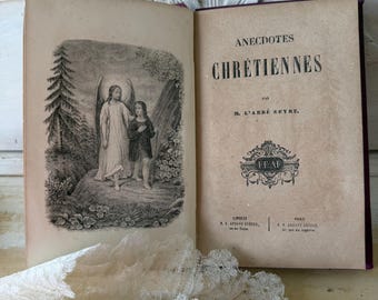 Puede incluir: Libro antiguo abierto con el título "ANECDOTES CHRÉTIENNES" y una ilustración de un ángel y una persona. El libro tiene una portada y páginas en tonos sepia, con texto en francés. Un trozo de encaje blanco está en primer plano.