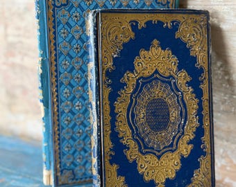 Puede incluir: Dos libros antiguos con cubiertas ornamentadas. Los libros tienen cubiertas azules con diseños dorados de filigrana. El lomo del libro de la izquierda es visible, mostrando una cubierta azul con un patrón floral. El libro de la derecha tiene un diseño dorado detallado.