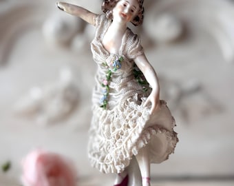 Puede incluir: Figura de porcelana de una mujer en pose de baile. La figura lleva un vestido de encaje blanco con detalles florales y zapatos burdeos. La base es blanca con ribete dorado. La mujer tiene el pelo castaño y está sonriendo.
