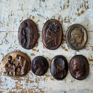 Könnte beinhalten: Eine Sammlung von sieben ovalen Wandplaketten in Bronzetönen mit klassischen Reliefs von Figuren und Profilen. Die Plaketten zeigen verschiedene Szenen, darunter sitzende Figuren, einen behelmten Kopf und individuelle Porträts im Relief.