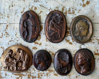Puede incluir: Una colección de siete placas de pared ovaladas en tonos bronce con relieves clásicos de figuras y perfiles. Las placas muestran varias escenas, incluyendo figuras sentadas, una cabeza con casco y retratos individuales en relieve.