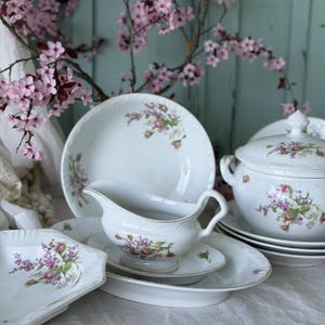 Könnte beinhalten: Ein weißes Porzellanservice mit floralen Designs in Rosa, Lila und Grün. Das Set umfasst Teller, eine Sauciere und eine Servierschüssel mit Deckel. Goldene Verzierungen akzentuieren die Ränder der Stücke und schaffen eine elegante Ästhetik.
