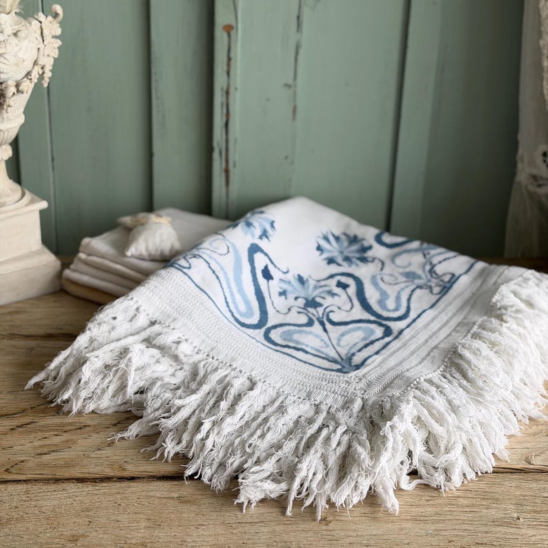 Puede incluir: Textil cuadrado blanco con un dise&ntilde;o floral azul y flecos. El dise&ntilde;o presenta flores azules estilizadas y l&iacute;neas fluidas. El textil tiene un grueso fleco en los bordes, a&ntilde;adiendo un toque decorativo.