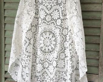Puede incluir: Una prenda de encaje blanco con un patrón floral. El intrincado diseño presenta varios motivos de flores y hojas, creando una textura delicada y aireada. La prenda parece ser un chal o una envoltura, con una forma fluida y drapeada.