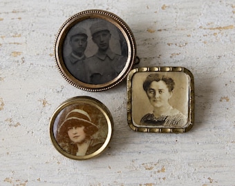 Puede incluir: Tres broches de metal de estilo antiguo con retratos fotográficos en blanco y negro. Un broche circular grande muestra a dos personas con gorras militares, uno circular más pequeño muestra a una persona con sombrero y uno cuadrado muestra un retrato.