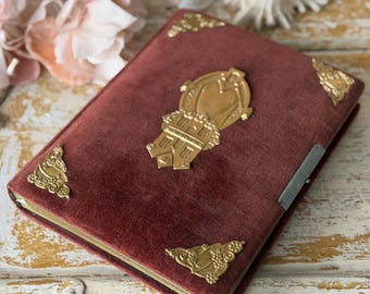 Puede incluir: Un álbum de fotos de terciopelo burdeos con esquinas de metal dorado y un emblema decorativo central. El álbum tiene un cierre de metal y descansa sobre una superficie de madera blanca envejecida, con conchas marinas y flores en el fondo.