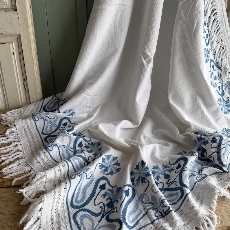Puede incluir: Manta blanca con borde de patr&oacute;n floral y remolino azul y flecos. La manta est&aacute; drapeada, mostrando el dise&ntilde;o detallado y la textura. El patr&oacute;n azul contrasta con el fondo blanco.