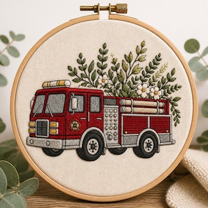 Puede incluir: Obra de arte bordada en un aro de madera que presenta un camión de bomberos rojo con ruedas negras y detalles plateados. El camión está adornado con un arreglo floral de hojas verdes y flores blancas. El fondo es de tela beige.