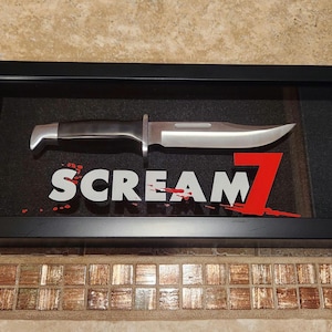 Könnte beinhalten: Eine schwarze, gerahmte Schattenbox mit einem silbernen Messer und dem Text "SCREAM 7" in weißen und roten Buchstaben.