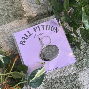 May include: A keychain with a silver ring and a pendant with a black and grey pattern. The keychain is on a purple card with the text "BALL PYTHON Mojave" and "BETHAN W ART".
