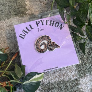 May include: A pin depicting a ball python in brown, tan, and white, on a light purple card. The card reads "BALL PYTHON Fire" and "BETHAN W ART". The pin is packaged in a clear plastic sleeve.