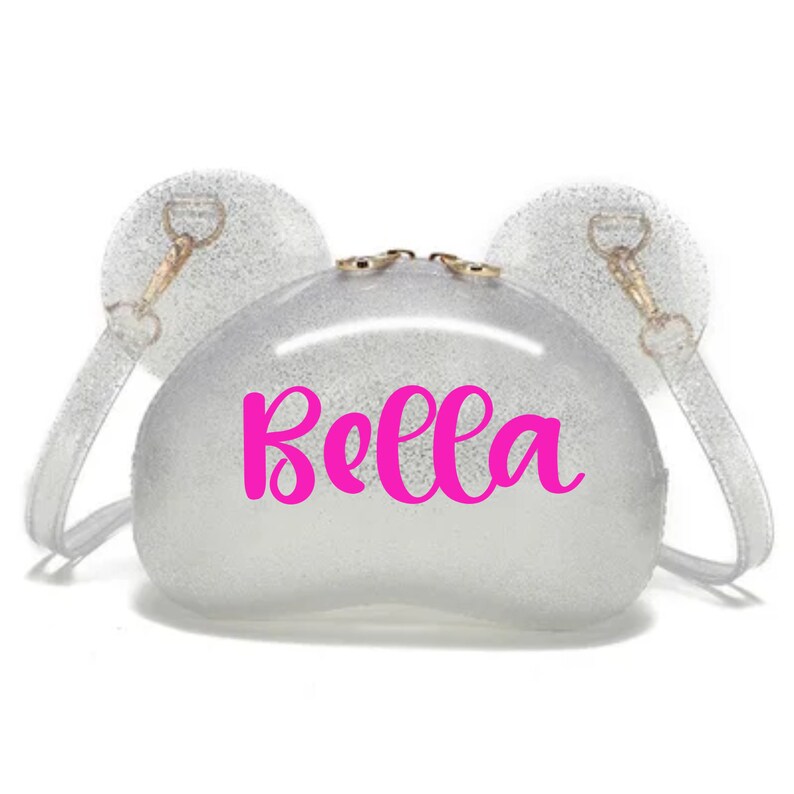 Puede incluir: Un bolso de pl&aacute;stico transparente con purpurina incrustada en el pl&aacute;stico. El bolso tiene dos asas en forma de orejas y la palabra "Bella" escrita en rosa en la parte delantera.