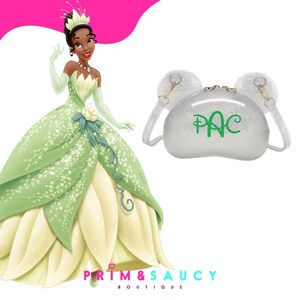 Puede incluir: Un vestido de princesa verde y blanco con un dise&ntilde;o de flores verdes. El vestido lo lleva una princesa de dibujos animados con piel oscura y una corona. Un bolso de purpurina plateado con un monograma verde "PAC" y dos asas con forma de orejas est&aacute; en el fondo.