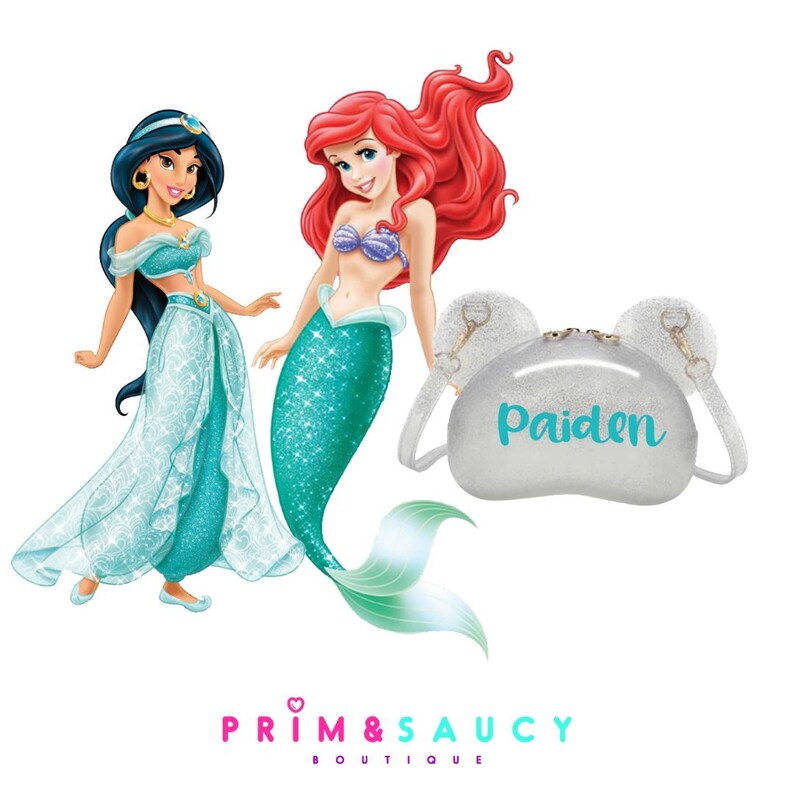Puede incluir: Un bolso de purpurina plateado con el nombre "Paiden" en azul turquesa en la parte delantera. El bolso tiene dos peque&ntilde;as orejas en la parte superior. El bolso est&aacute; al lado de una imagen de dibujos animados de Ariel de La Sirenita y Jasmine de Aladdin.