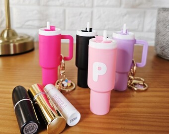 May include: A collection of colorful tumbler-shaped keychains in pink, black, and purple, with a white lid and handle. The pink keychain has the letter "P" on it. Also, there are three lipsticks and a lip balm on the wooden surface.
