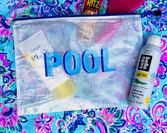 Puede incluir: Una bolsa de malla blanca con cremallera con la palabra "POOL" en letras azules. La bolsa está llena de varios artículos, incluyendo una botella de protector solar, una caja de galletas y un cepillo de pelo rosa. La bolsa está sentada sobre una tela de estampado floral azul y rosa.