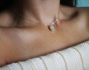 May include: A delicate gold chain necklace with a small, oval-shaped pendant featuring a sparkly, white crystal design.