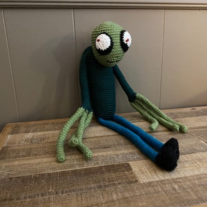 May include: Green crocheted plush toy with large eyes and a long, thin body. The toy is sitting on a wooden surface.