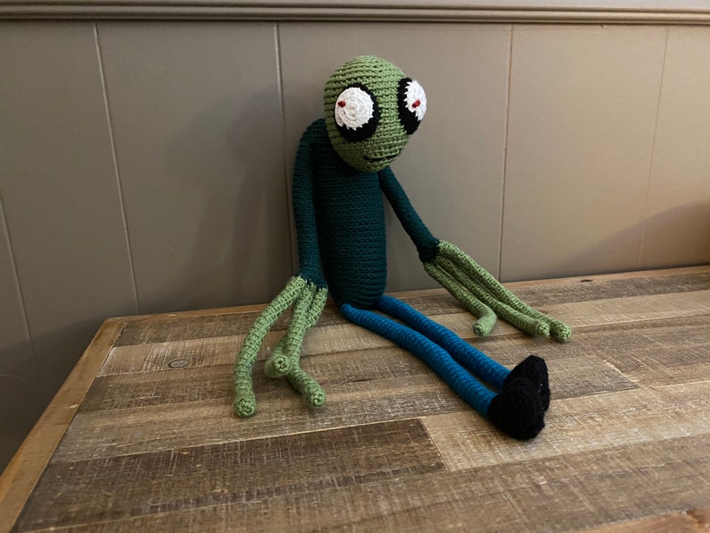 May include: Green crocheted plush toy with large eyes and a long, thin body. The toy is sitting on a wooden surface.
